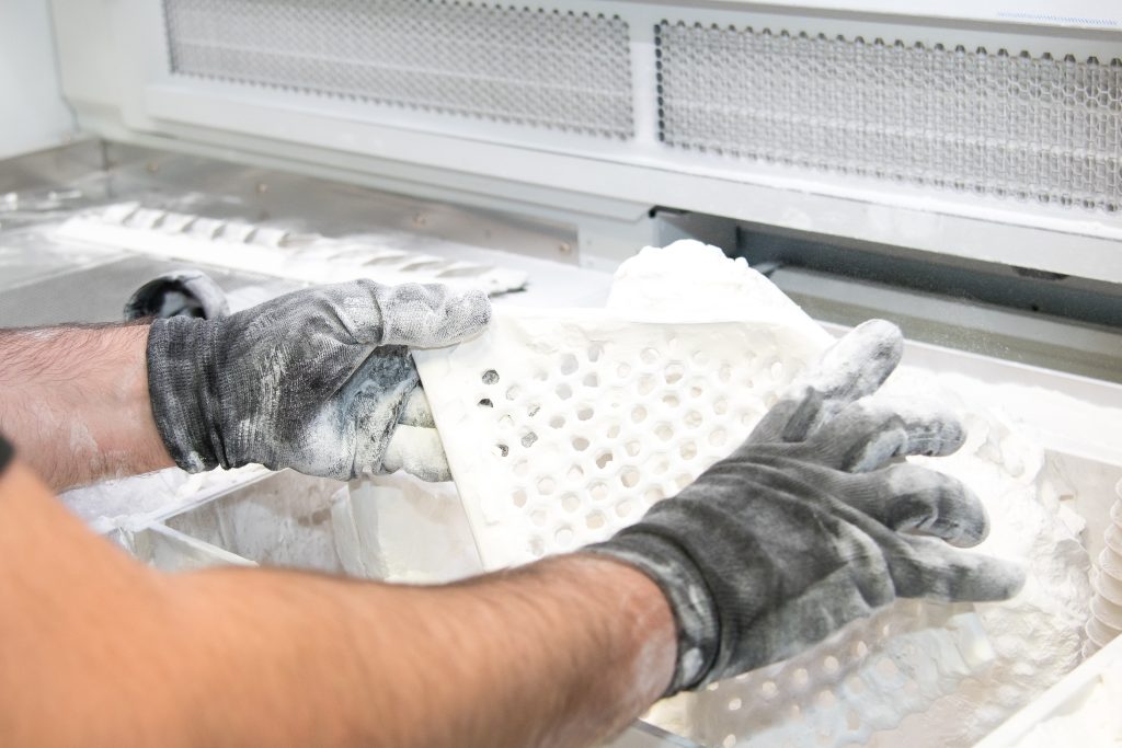 A pair of hands wearing gloves working with a 3d printed part. The component is being covered in a white powder as part of the production process.