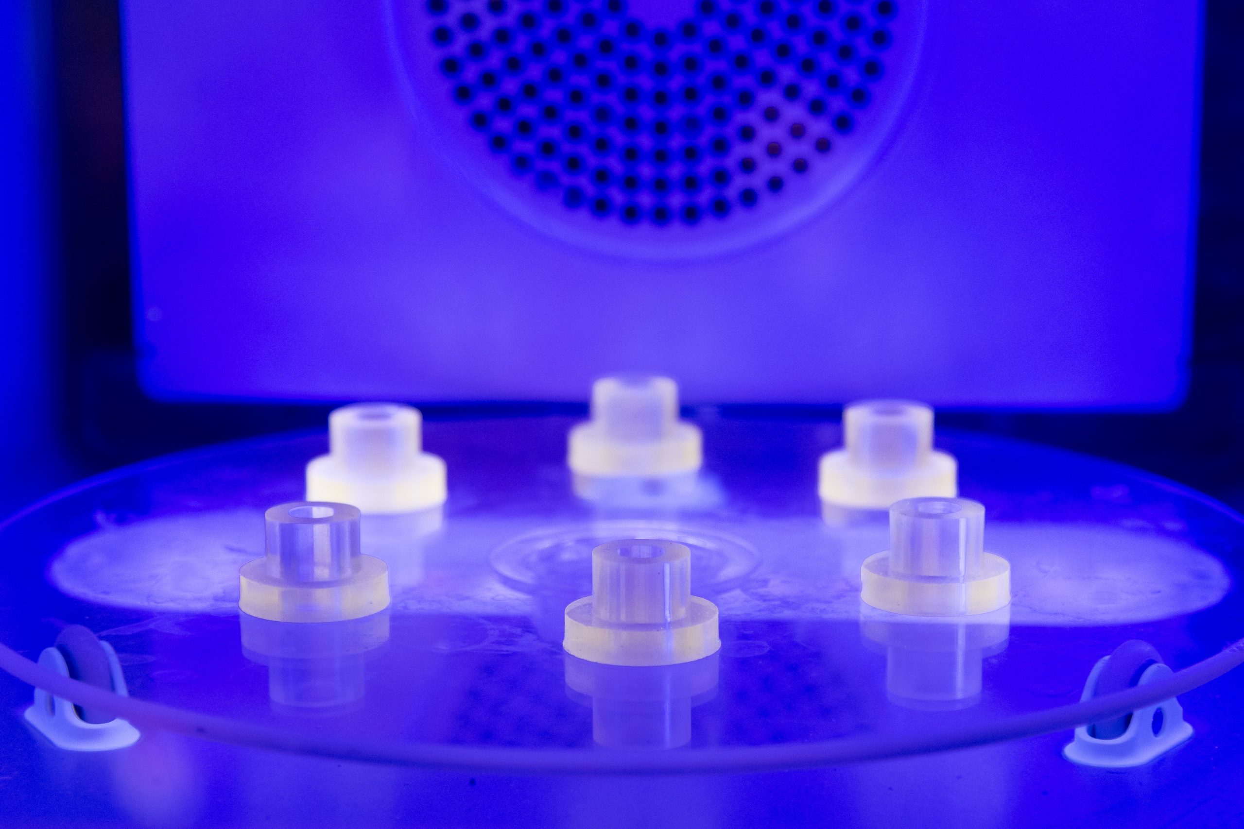 The inside of a 3d printer. The centre of the machine has six cylinders and is lit by a deep blue light.