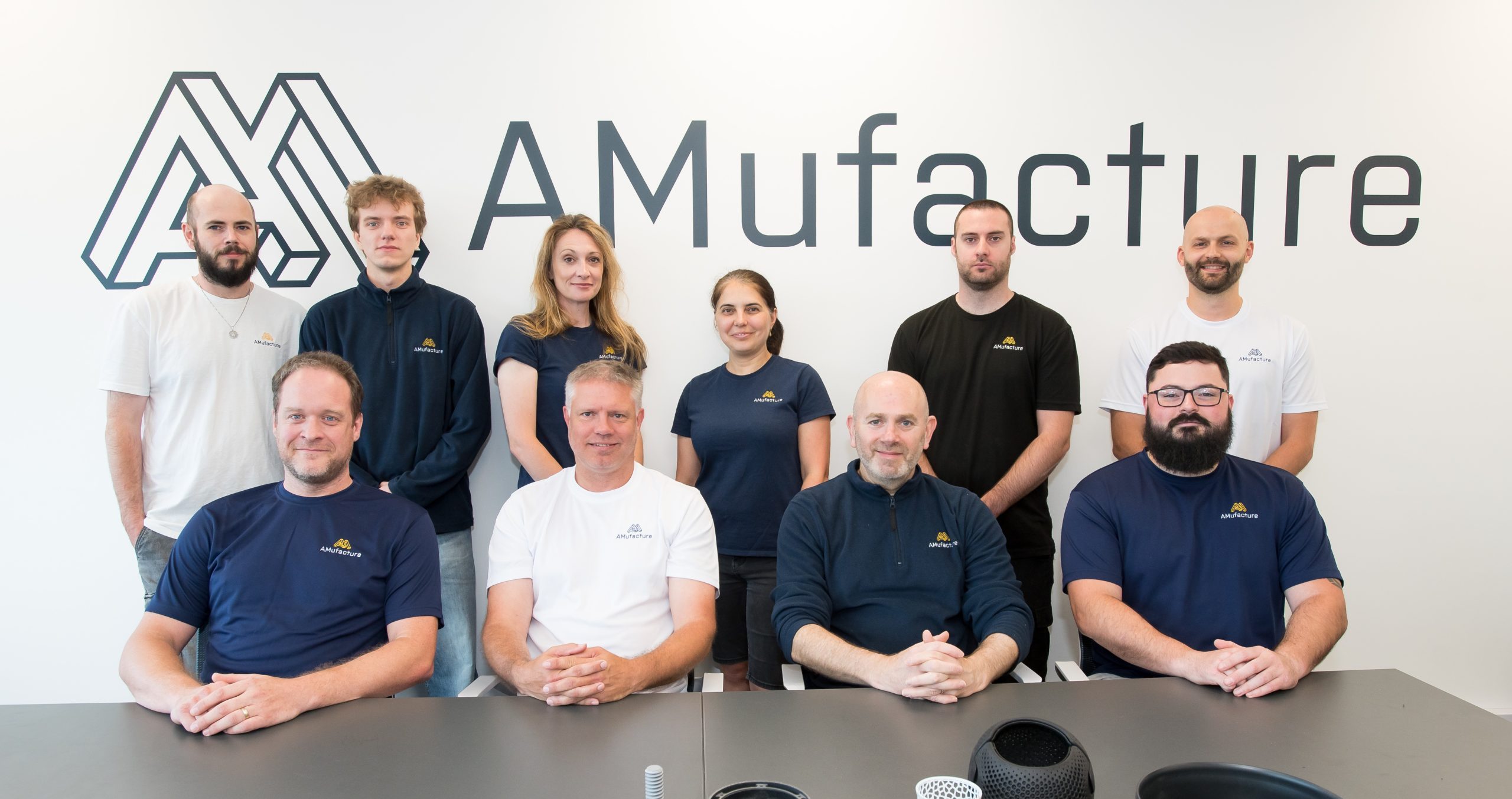 A group of ten people, four are sat at a table and six are stood behind them. There is a collection of different 3D printed components on the table.