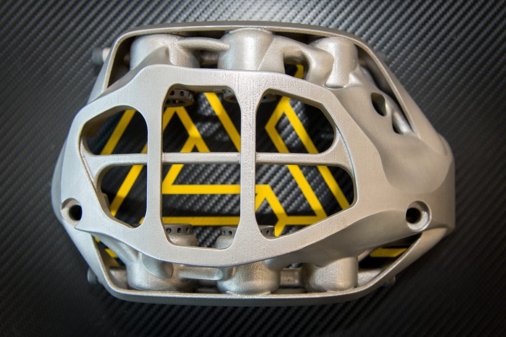 A 3D printed automotive part with a yellow AMufacture logo behind it.